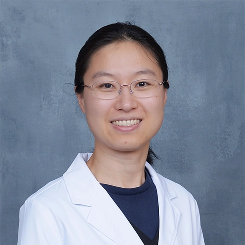 Interventional Pain Physician Danqing Hu In Lab Coat Smiling In Front Of Blue Background