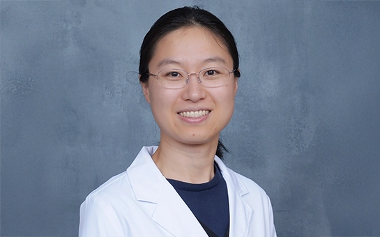Interventional Pain Physician Danqing Hu Smiling In Lab Coat In Front Of Blue Background